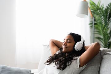 Indian woman relaxing sitting on sofa with copy space. Teenager listening to music on headphones.