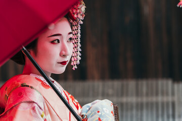 A contemplative geisha in a striking red kimono holds a parasol, her gaze introspective amidst a traditional setting. Her visage tells a story of cultural depth