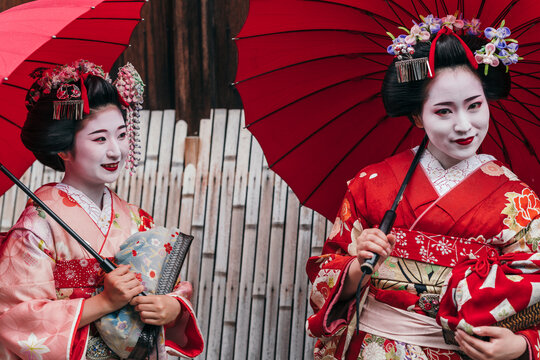 Two maiko geishas share a light-hearted moment, their red umbrellas adding a pop of color against their ornate kimonos. Their camaraderie shines through the cultural attire