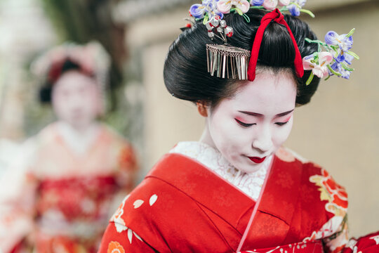 A geisha in a vibrant red kimono contemplates quietly, her face a study of elegance. Her elaborate hair ornaments signal her status in Japanese cultural tradition
