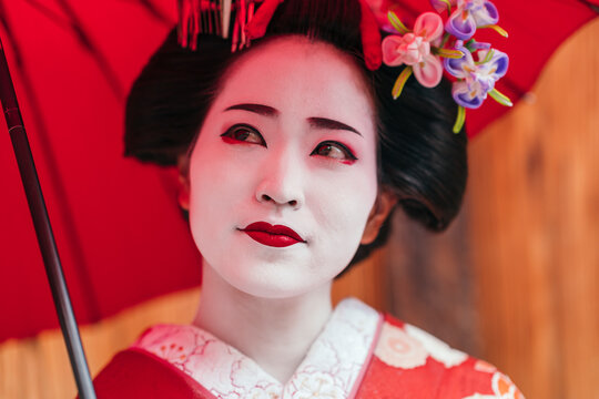 The thoughtful expression of a geisha in a vibrant red kimono captures a moment of calm, her red parasol providing a striking contrast. The image is a study in composed beauty.