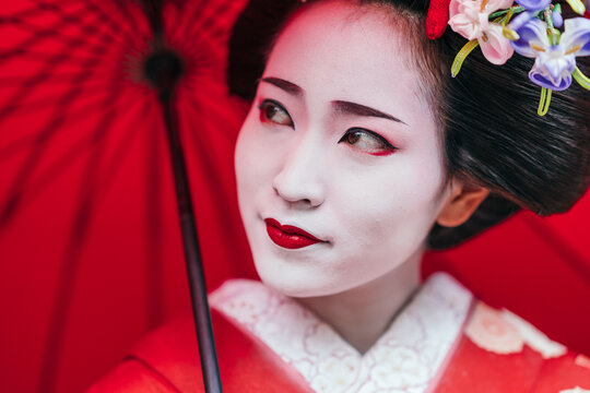 A geisha's contemplative gaze peeks from beneath her red umbrella, surrounded by the textures of her traditional attire. The image evokes a sense of quiet introspection.