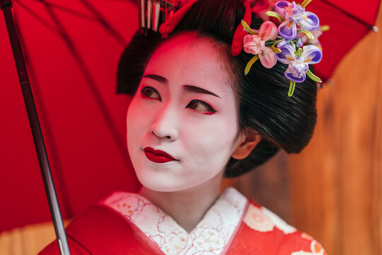 A geisha adorned with vibrant red lips and an umbrella gazes serenely. Traditional hair ornaments and red accents complement her striking look