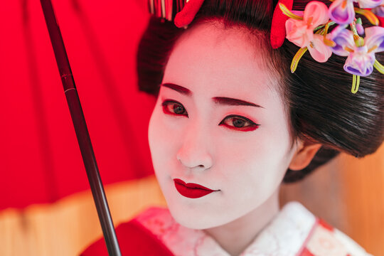 A geisha's gentle smile peeks beneath a red umbrella, her joyful aura complemented by the ornate kimono. The red and white contrast paints a classic image of Japanese beauty