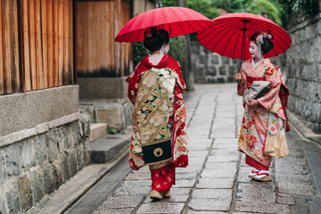A pair of Geishas in elaborate kimonos walk under red umbrellas on a cobblestone path, a perfect scene of Kyoto's tradition