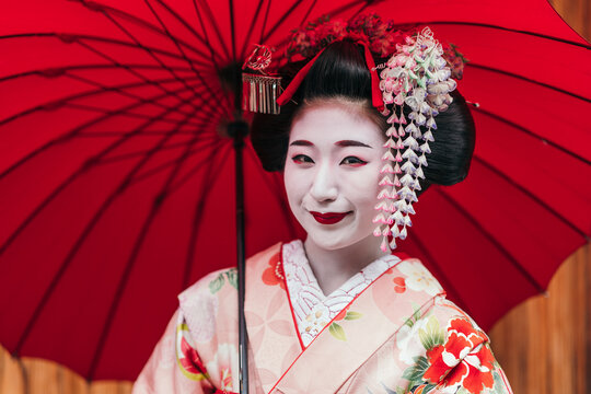 A smiling Geisha under a red parasol, exuding elegance in her red and white patterned kimono, embodies the grace of Japanese culture