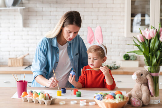 Cheerful mother with her little son painting eggs for Easter in the kitchen at home. Young happy family of two decorating ornaments for holiday celebration together