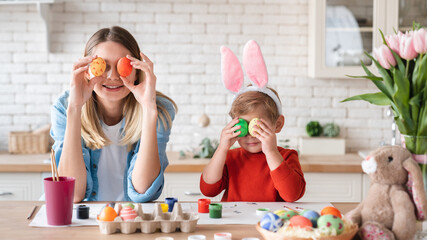 Playful mom and little son holding Easter eggs close to their eyes in the kitchen. Decorations for...