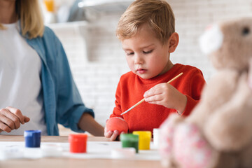 Close up portrait of a little boy painting Easter eggs with his mom at home. Family time and new...