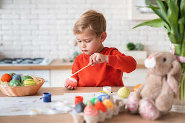 Small european boy decorating eggs for Easter in the kitchen. Caucasian little child kid son playing painting making holiday decorations for celebration at home