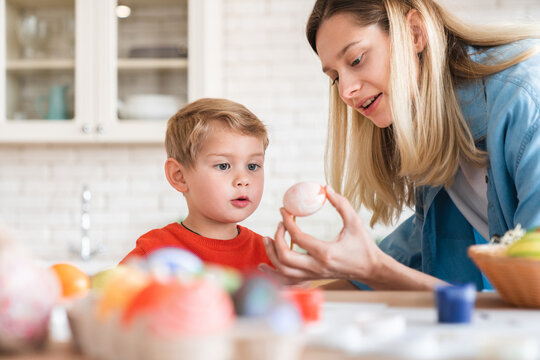 Young caucasian mom helps her little son decorate eggs for Easter at home. Mother and happy small kid child painting decorations ornaments, spending time and preparing for celebration