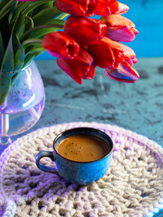 Coffee cup on a knitted napkin. A bouquet of red tulips in a vase. Blue background