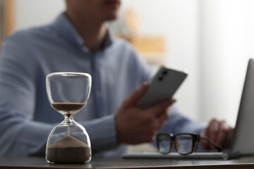 Hourglass with flowing sand on desk. Man using smartphone while working on laptop indoors, selective focus - obrazy, fototapety, plakaty