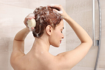 Young woman washing her hair with solid shampoo bar in shower, back view