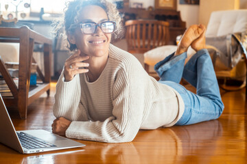 Side view of cheerful young adult woman using laptop at home laying on the floor and smiling...