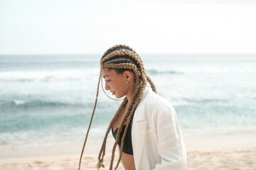 Portrait of a young Latino woman with braids on her head. She walks along the beach near the ocean in a jacket and suit. Self confidence and masculinity concept. Braiding studio.