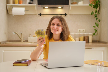 A woman with a laptop holds euro money at a table in a home kitchen. An adult female businesswoman...