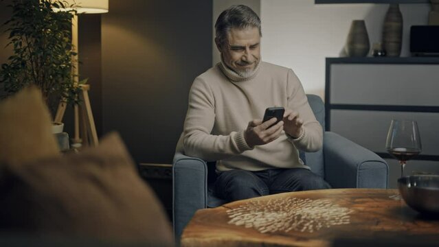 Older man sitting in cosy living room at home using cell phone, reading email, checking social media, playing, working online with smartphone app. Focused mid adult male.