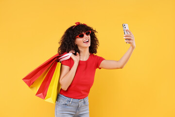 Happy young woman with shopping bags taking selfie on yellow background