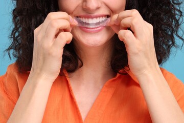 Young woman applying whitening strip on her teeth against light blue background, closeup