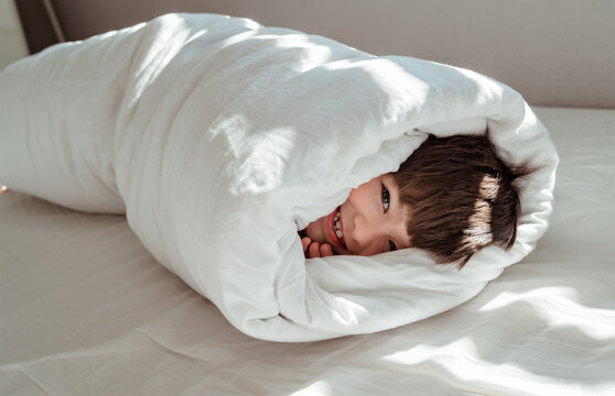Little happy toddler boy wrapped in blanket like a roll looking at camera smiling having fun on the bed.