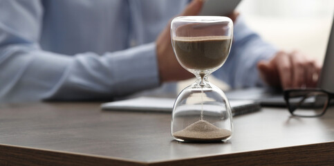 Hourglass with flowing sand on desk. Man using smartphone while working on laptop indoors,...
