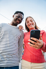 Vertical portrait of two teenage happy friends watching funny videos browsing on internet app with a smartphone and smiling together. Multiracial couple using a cell phone to post in a social media