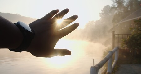 A person's hand is holding up to the sun, with a body of water in the background
