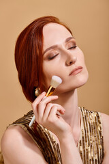 redhead woman in gold earring applying face foundation with makeup brush on beige backdrop