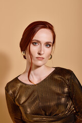 attractive redhead woman in 30s posing in stylish outfit and golden accessories on beige backdrop