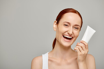 Redhead young and cheerful woman in 30s holding tube with body lotion on grey background, skincare