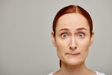 redhead and worried woman in white tank top embodying concern on grey background, concerned face
