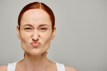 redhead young woman in white tank top frowning and grimacing on grey background, concerned face