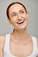 happy redhead woman in tank top exuding joyful and healthy smile on grey background, looking away