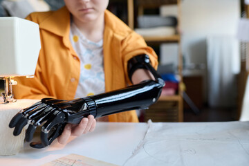 Close up of young woman with disability adjusting prosthetic arm while using sewing machine copy...
