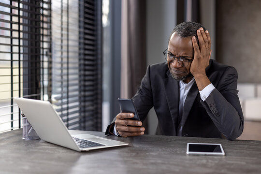 Stressed african american entrepreneur grabbing head with hand while using smartphone in remote workspace. Frightened man getting warning notification from banking application while sitting by desk
