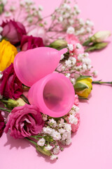 Menstrual cups beside a collection of vibrant, fresh flowers on a pink background, depicting an...