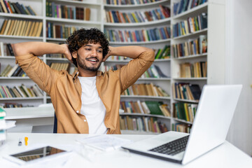Positive hispanic guy leaning on back of chair while holding hands behind head and looking at...