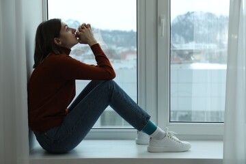 Sad young woman sitting on windowsill near window at home, space for text