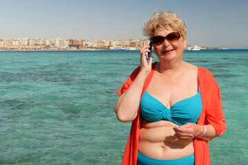 Senior adventure is ageless plus size woman wearing in swimsuit speak by mobile phone on the beach. Summer vacation for pensioner.