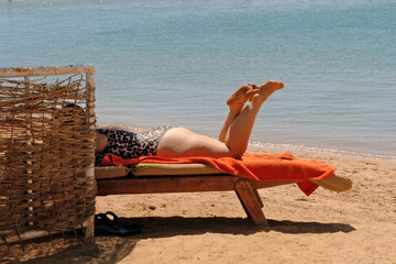 unknown woman lies on a wooden deck chair and enjoys rest and relaxation on the beach against the backdrop of the sea on a sunny summer day, copy space