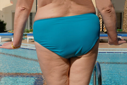 an overweight woman in a swimsuit with cellulite stands with her back and does fitness near the pool before swimming in the summer on a sunny day, outdoors,