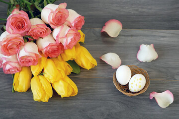 Many yellow tulips and pink roses bouquet and eggs in bowl lying at grey wooden background, happy easter concept