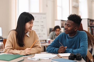 Side view portrait of smiling Asian woman looking at Black student while studying together in college library