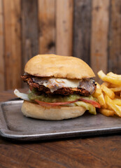 Fast food. Closeup view of a chicken burger with potato bread, cheese, lettuce, tomato, guacamole and french fries, in a metal dish on the wooden table.	
