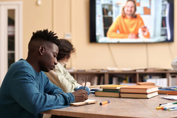 Side view portrait of Black young man taking notes during online lecture with teacher on screen in...