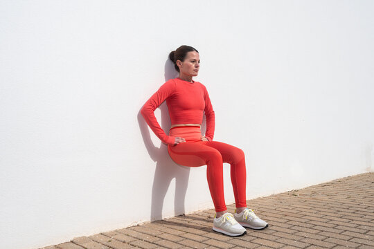Sporty woman doing squats against a white wall outside in the sun.