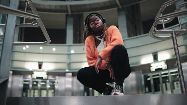 Stylish black female with vitiligo skin enjoying song beat in wireless headphones. Attractive African American listen to music from online playlist through earbuds while sitting in subway. Slow motion