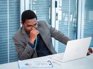 A handsome African American businessman in a suit is sitting at a work desk analyzing financial data and doing research