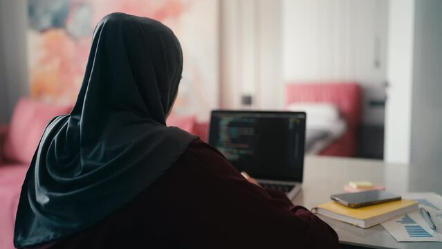 Young woman in hijab testing software code on laptop, working as IT specialist